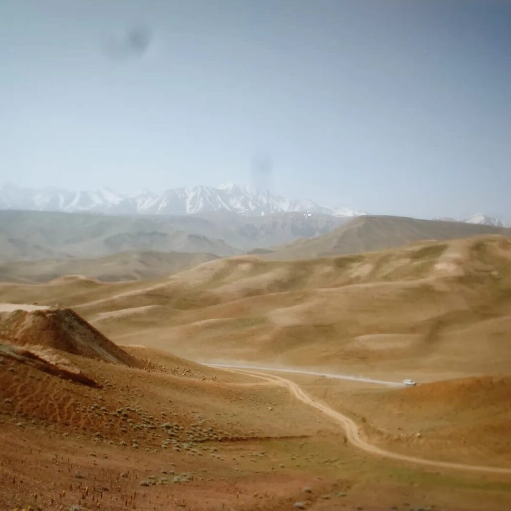 Weite Berglandschaft in Bamiyan mit schneebedeckten Gipfeln im Hintergrund und einer Schotterstraße im Tal