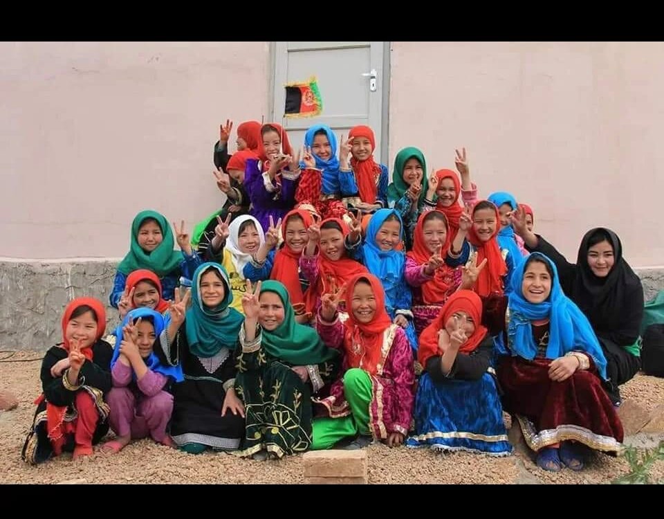 Große Gruppe von Mädchen und jungen Frauen in bunten Kleidern zeigt Victory-Zeichen vor einem Gebäude mit afghanischer Flagge