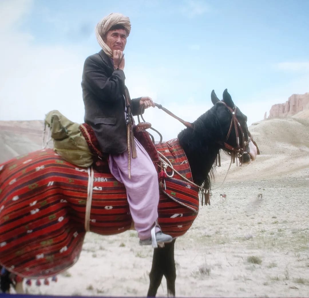 Mann mit Turban reitet auf einem geschmückten Pferd durch eine trockene Landschaft in Bamiyan